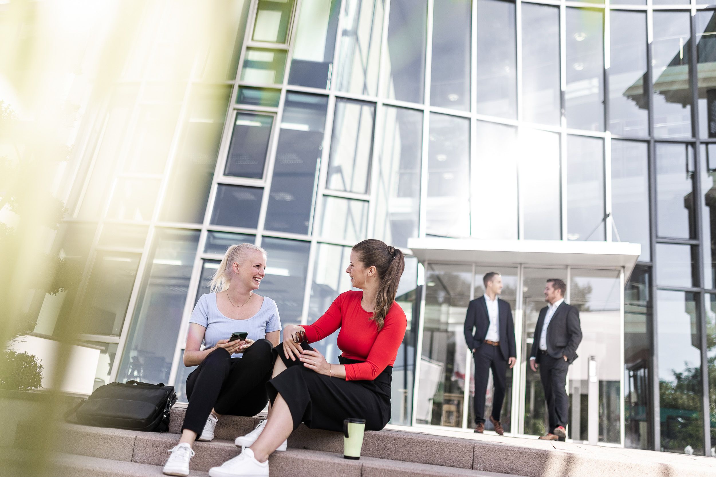 Content Creation Frankfurt bei Werbefotografie Shooting - 2 Mitarbeiterinnen sitzen locker vor Unternehmen im Hintergrund zwei Männer und ein Glasgebäude