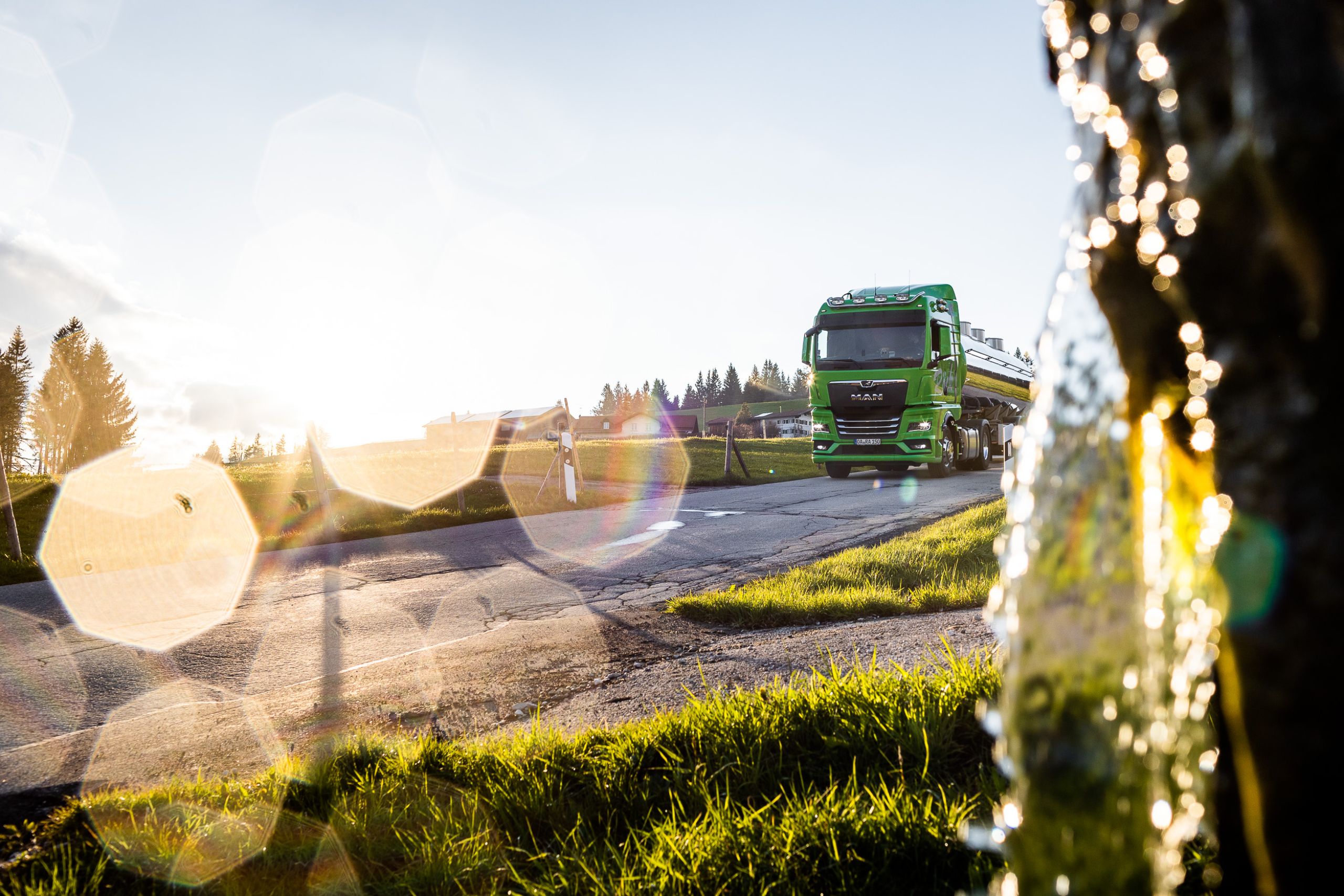 Werbefotografie für Nutzfahrzeughersteller - MAN TGX mit Milch Tank im Allgäu fährt auf einer Straße an einem Brunnen vorbei im Sonnenuntergang